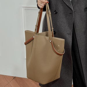 May include: A tan leather tote bag with brown handle detailing. The bag has a rectangular shape with a spacious interior and long shoulder straps. The handles are a darker brown colour, and the bag appears to be made of soft, textured leather.