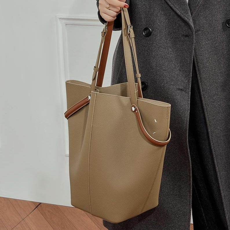 May include: A tan leather tote bag with brown handle detailing. The bag has a rectangular shape with a spacious interior and long shoulder straps. The handles are a darker brown colour, and the bag appears to be made of soft, textured leather.