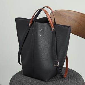 May include: Black leather tote bag with brown leather handles and a detachable shoulder strap. The bag has a structured design with a spacious interior. The handles are attached with silver-tone hardware. The bag is sitting on a grey chair.