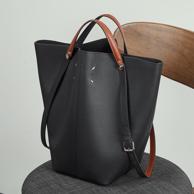 May include: Black leather tote bag with brown leather handles and a detachable shoulder strap. The bag has a structured design with a spacious interior. The handles are attached with silver-tone hardware. The bag is sitting on a grey chair.