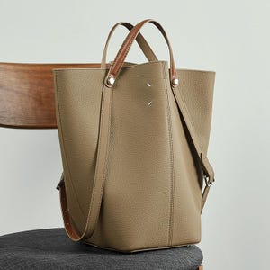 May include: A taupe tote bag with brown leather handles and a detachable shoulder strap. The bag has a rectangular shape with a textured surface and silver-tone hardware. The handles are attached with silver rivets.