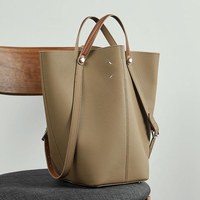 May include: A taupe tote bag with brown leather handles and a detachable shoulder strap. The bag has a rectangular shape with a textured surface and silver-tone hardware. The handles are attached with silver rivets.