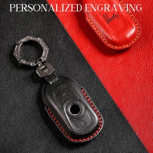 Puede incluir: Funda de llavero de cuero negro con costuras rojas y un llavero de metal plateado. La funda tiene una abertura circular y un diseño en relieve. También se ve una funda de llavero de cuero rojo con la palabra "liberty".