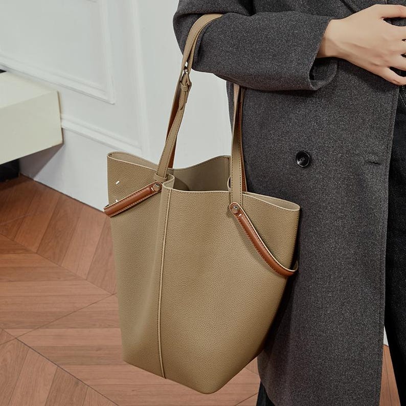 May include: A tan leather tote bag with brown handles and accents. The bag has a structured design with a spacious interior and long shoulder straps. The bag is being held by a person wearing a grey coat.