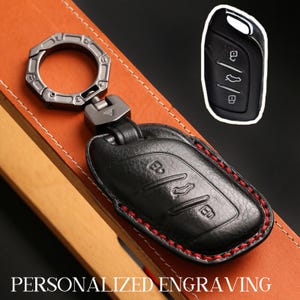 Puede incluir: Funda de llave de coche de cuero negro con costuras rojas, sujeta a un llavero de metal plateado. El llavero tiene tres botones y se muestra sobre una superficie de cuero marrón. El texto "PERSONALIZED ENGRAVING" está en la parte inferior.
