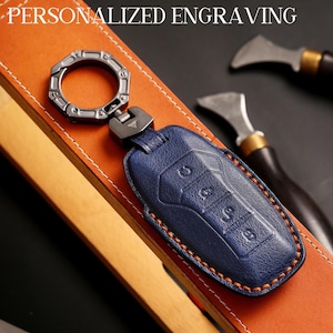 May include: A blue leather key fob with orange stitching and a metal key ring. The key fob has buttons and is attached to a metal clasp. The words "PERSONALIZED ENGRAVING" are at the top of the image.