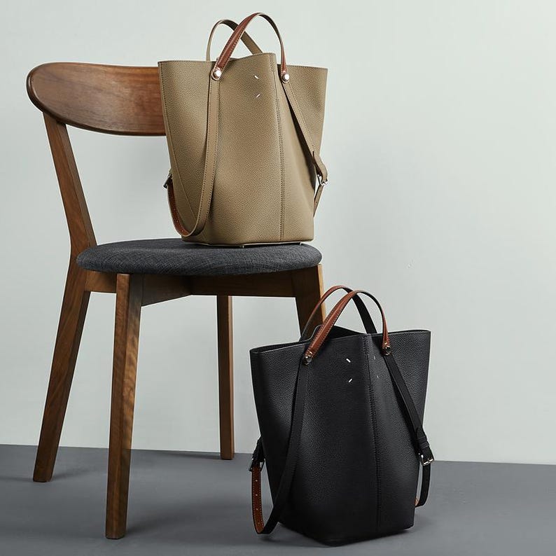 May include: Two leather tote bags, one in tan and one in black, with brown handles and shoulder straps. The bags are on a wooden chair with a grey seat. The bags have a minimalist design.