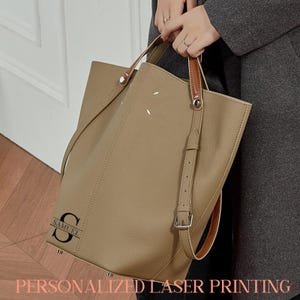 May include: A beige leather tote bag with brown leather handles and a detachable shoulder strap. The bag has a personalized laser-printed initial and name. The text "PERSONALIZED LASER PRINTING" is displayed at the bottom of the image.
