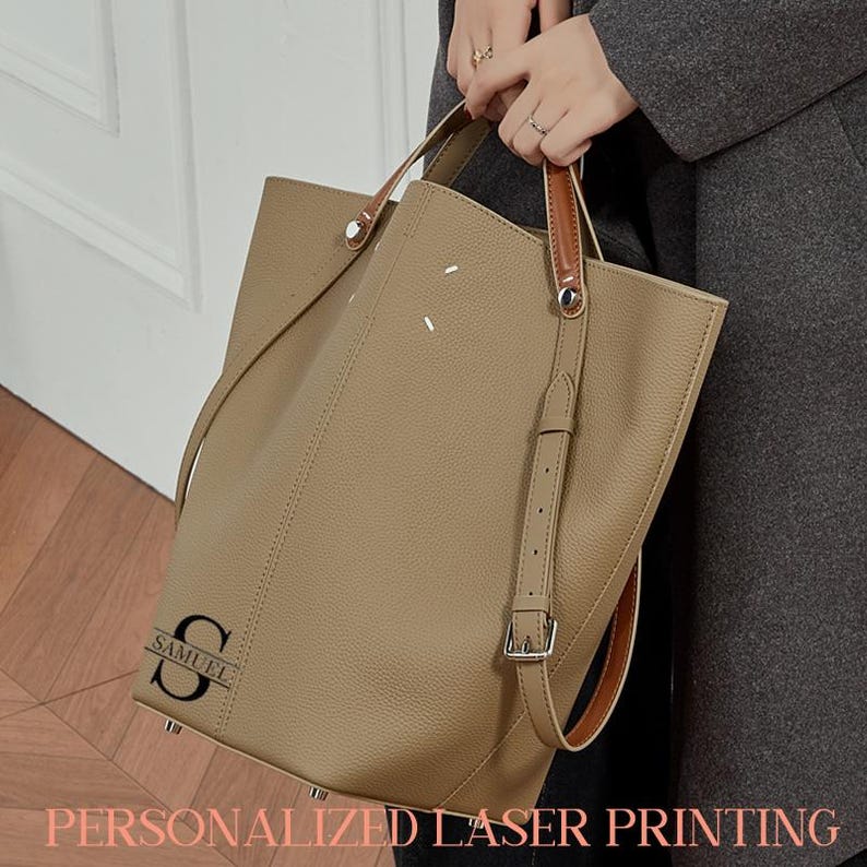 May include: A beige leather tote bag with brown leather handles and a detachable shoulder strap. The bag has a personalized laser-printed initial and name. The text "PERSONALIZED LASER PRINTING" is displayed at the bottom of the image.