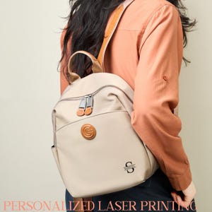 Puede incluir: Mochila beige con detalles de cuero marrón, un bolsillo frontal y cremalleras plateadas. La mochila tiene un parche de cuero circular y una inicial y nombre personalizados. El texto "PERSONALIZED LASER PRINTING" está en la parte inferior.