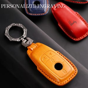 May include: An orange leather key fob cover with a silver key ring and clasp. The cover has embossed buttons and stitching around the edges. The text "PERSONALIZED ENGRAVING" is at the top. Other colored key fob covers are in the background.