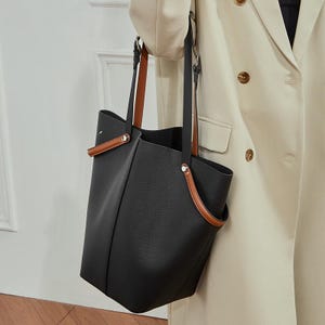 May include: A black tote bag with brown leather handles and trim. The bag has a structured shape and a minimalist design. The handles are attached with silver-coloured hardware. The bag is held by a person wearing a cream-coloured blazer.