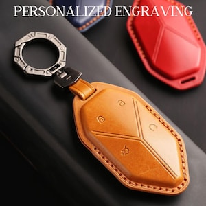 May include: A tan leather key fob with a silver metal ring and clasp. The fob features a geometric design with embossed symbols. The words "PERSONALIZED ENGRAVING" are at the top. A red and blue key fob are in the background.