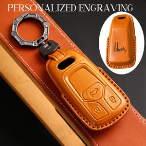 May include: A tan leather key fob cover with embossed key symbols and a metal key ring. The cover has a stitched border and is shown on a leather strap. The text "PERSONALIZED ENGRAVING" is at the top.