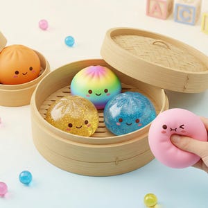 May include: A collection of colorful, smiling dumpling-shaped stress balls in bamboo steamers. The dumplings are orange, rainbow, gold glitter, and blue glitter. A pink dumpling is being squeezed by a hand. Scattered around are small, colorful, spherical beads.
