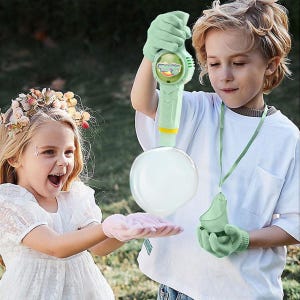 May include: Two children playing with a bubble gun. The bubble gun is green and yellow. One child is wearing green gloves and holding the bubble gun. The other child is wearing pink gloves and reaching for a large bubble.