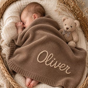 May include: A sleeping baby wrapped in a brown knitted blanket with the name "Oliver" embroidered in cream-colored thread. A teddy bear with a checkered bow tie sits beside the baby in a woven basket. The blanket has a ribbed border.