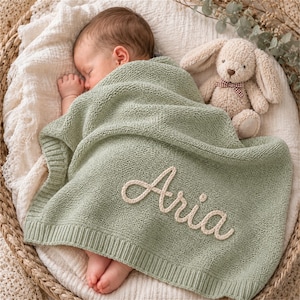 May include: A baby rests peacefully under a sage green knitted blanket embroidered with the name "Aria" in cream-colored thread. A stuffed bunny with a red and white checkered bow tie sits beside the baby in a woven basket.