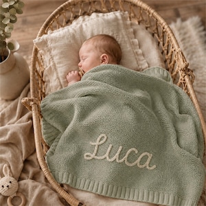 May include: A sleeping baby is nestled in a wicker bassinet, covered with a sage green knitted blanket. The blanket is embroidered with the name "Luca" in a cream-colored script. A cream-colored pillow supports the baby's head.