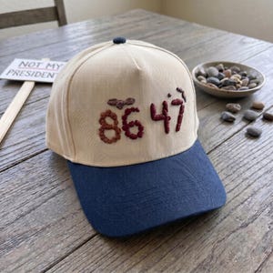 Op de afbeelding: Een beige en marineblauwe baseballpet met de geborduurde cijfers "86 47" in bordeauxrood garen. De pet heeft een donkerblauwe knoop bovenop en een gebogen klep. Een bord met de tekst "NOT MY PRESIDENT" staat op de achtergrond.