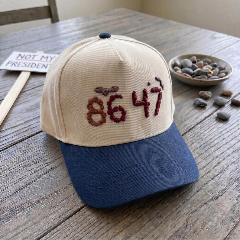 Op de afbeelding: Een beige en marineblauwe baseballpet met de geborduurde cijfers "86 47" in bordeauxrood garen. De pet heeft een donkerblauwe knoop bovenop en een gebogen klep. Een bord met de tekst "NOT MY PRESIDENT" staat op de achtergrond.