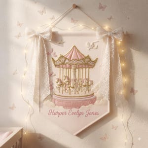 May include: A cream-colored fabric wall banner featuring a carousel illustration with pink and gold accents, adorned with two large white lace bows at the top corners. The name Harper Evelyn Jones is printed in pink script at the bottom of the banner.