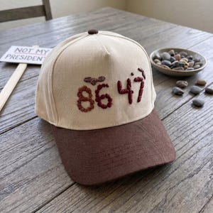 Op de afbeelding: Een baseballpet met een cr&egrave;mekleurige kroon en een bruin vizier. De pet heeft geborduurde kastanjebruine cijfers "86 47". Bovenop de pet zit een kleine bruine knoop. Op de achtergrond staat een bord met "NOT MY PRESIDENT".