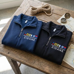 May include: Two navy and black quarter-zip sweatshirts with the name "ZIDANE" embroidered in white, along with nautical flag designs. The sweatshirts are displayed on a wooden table with nautical accessories, including a rope and a compass.