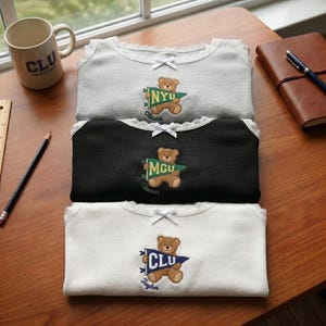 May include: Three ribbed shirts in gray, black, and white, each featuring a lace-trimmed neckline with a small white bow. Each shirt displays an embroidered brown teddy bear holding a pennant flag with the text NYU, MGU, or CLU and a name below.