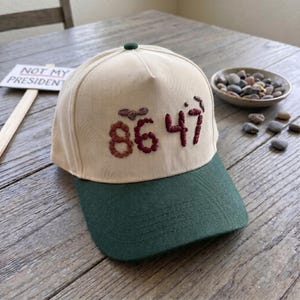 Op de afbeelding: Een baseballpet met een cr&egrave;mekleurige kroon en een donkergroene klep. De pet heeft geborduurde bordeauxrode cijfers "86 47". Een klein bordje met "NOT MY PRESIDENT" staat op de achtergrond.