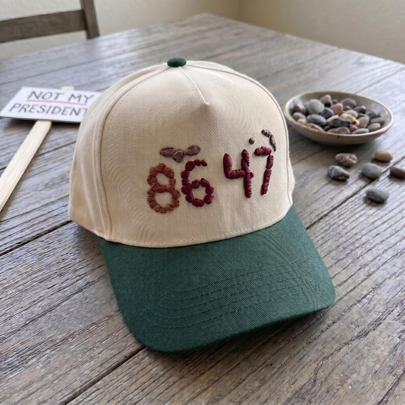 Op de afbeelding: Een baseballpet met een cr&egrave;mekleurige kroon en een donkergroene klep. De pet heeft geborduurde bordeauxrode cijfers "86 47". Een klein bordje met "NOT MY PRESIDENT" staat op de achtergrond.