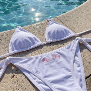May include: White bikini set with triangle top and tie sides. The bottom features the embroidered text "Mrs. Lily" in pink script. The swimwear is displayed near a pool.