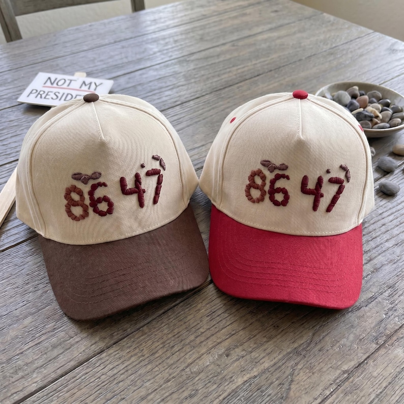 Op de afbeelding: Twee baseballpetten met beige kronen en contrasterende klepjes. E&eacute;n heeft een bruin klepje, de andere een rood klepje. Beide petten hebben het geborduurde nummer "86 47" in donkerbruin. De tekst "NOT MY PRESIDENT" is zichtbaar op de achtergrond.
