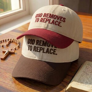 May include: Two baseball caps stacked on top of each other. The top cap is beige with a maroon brim and the text "GOD REMOVES TO REPLACE" embroidered in maroon. The bottom cap is beige with a brown brim and the same text.