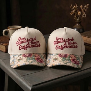 May include: Two cream-colored baseball caps with the embroidered text "Over Stimulated & Under Caffeinated" in burgundy. The brim features a floral pattern with red, pink, and green flowers. The caps are displayed on a dark gray surface.