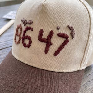 Op de afbeelding: Beige baseballpet met een bruine klep. De pet heeft geborduurde cijfers "8647" in een donkerroodbruine draad. De hoed is gemaakt van een zachte stof en heeft een gebogen klep.