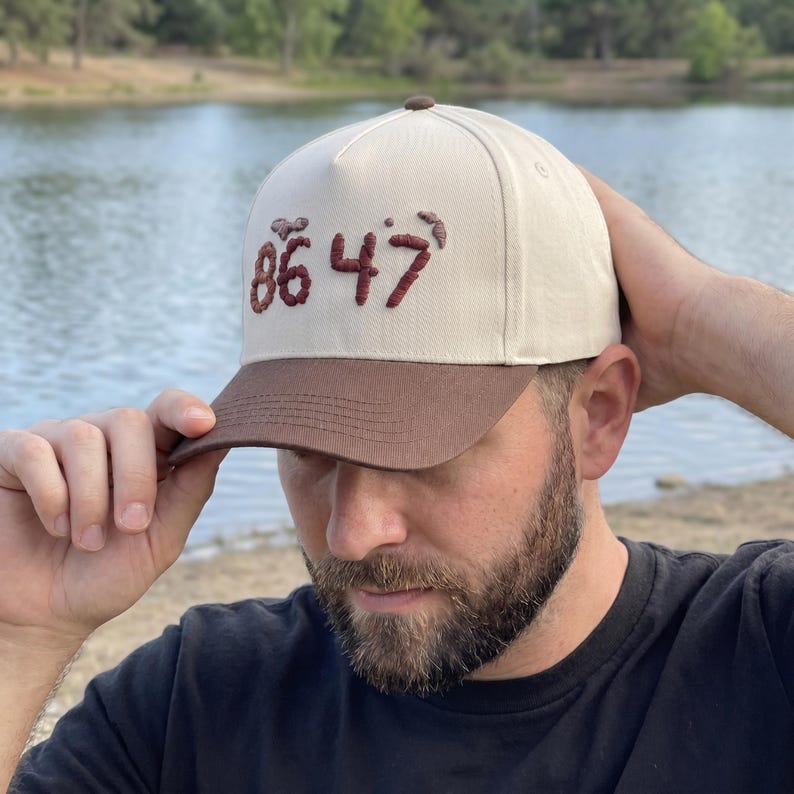 Op de afbeelding: Een beige en bruine baseballcap met de geborduurde cijfers "86 47" in donkerrood. De cap heeft een gebogen klep en een knoop aan de bovenkant. De achtergrond toont een meer en bomen.