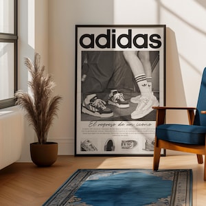 May include: A framed Adidas poster featuring a black and white image of sneakers and the Adidas logo. The poster is displayed in a room with a blue chair, a blue rug, and a potted plant.