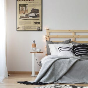 May include: A framed poster featuring a Nike X Travis Scott sneaker design hangs above a bed with gray bedding and decorative pillows. A small side table with a lamp and a rug with a black and white geometric pattern are in the foreground.