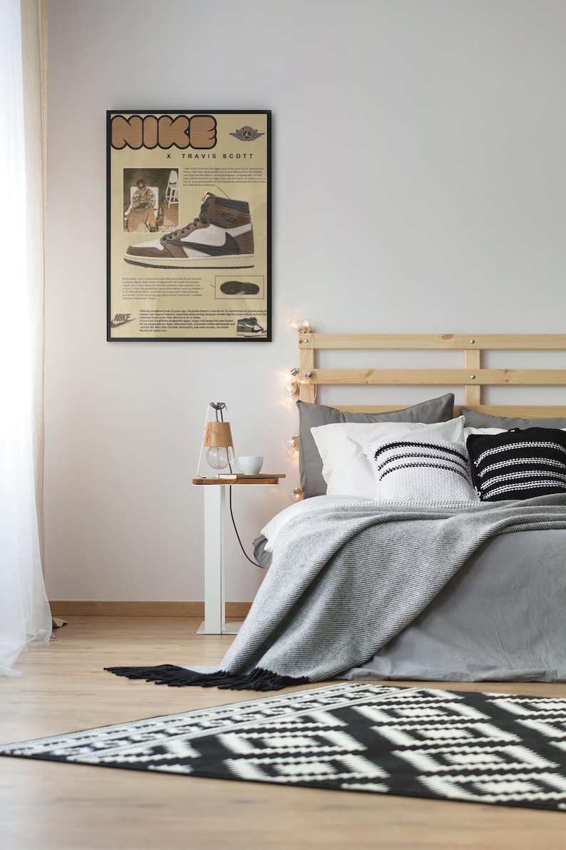 May include: A framed poster featuring a Nike X Travis Scott sneaker design hangs above a bed with gray bedding and decorative pillows. A small side table with a lamp and a rug with a black and white geometric pattern are in the foreground.