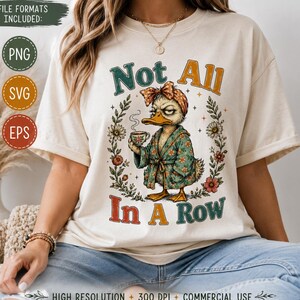 Pode incluir: Uma t-shirt cor de creme com o gráfico de um pato a usar um roupão floral e um lenço a condizer enquanto segura uma chávena de café. O texto Not All In A Row está rodeado por uma coroa de flores. A t-shirt tem mangas curtas.