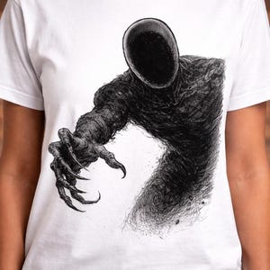 May include: White t-shirt featuring a detailed black and white illustration of a faceless, shadowy figure with outstretched arm and clawed hand. The artwork has a textured, pen-and-ink style.