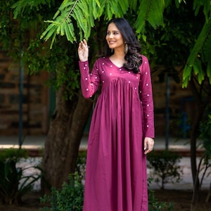 May include: A long, burgundy dress with a V-neck and long sleeves, adorned with small white polka dots. The dress has a gathered waist and is paired with matching burgundy pants. The outfit is worn outdoors, with a backdrop of green foliage and a tree.
