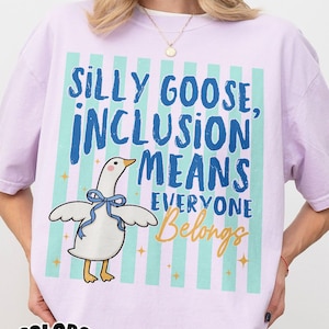 Puede incluir: Camiseta de color lila claro con las palabras "SILLY GOOSE, INCLUSION MEANS EVERYONE Belongs" en azul y naranja. Presenta un dibujo de un ganso con un lazo azul. La camiseta está etiquetada como "COMFORT COLORS Orchid".