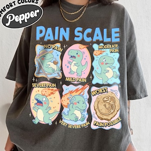 Op de afbeelding: Donkergrijs t-shirt met een kleurrijke afbeelding van een pijnscore met cartoon dinosaurussen. De tekst op het shirt luidt "PAIN SCALE" en bevat niveaus van "No Pain" tot "Pain Possible". Het shirt heeft ook de tekst "COMFORT COLORS Pepper".