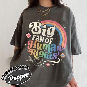 Peut inclure: Un t-shirt gris anthracite à manches courtes avec une impression graphique colorée indiquant Big Fan of Human Rights dans une police rétro. Le motif comprend un arc-en-ciel, des petites fleurs et une colombe tenant un rameau d'olivier.