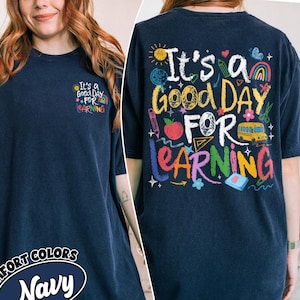May include: A navy blue short-sleeve t-shirt featuring a small graphic on the front left chest and a large graphic on the back. Both graphics display the text "It's a GOOD DAY FOR LEARNING" surrounded by colorful school-themed icons like a bus, apple, and rainbow.