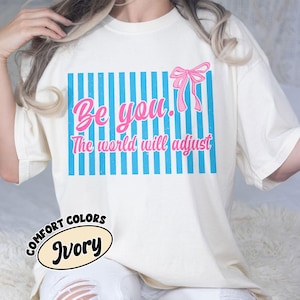 Puede incluir: Camiseta de color marfil con un diseño gráfico. El diseño presenta las palabras "Be you. The world will adjust" en rosa, sobre un fondo de rayas azules con un lazo rosa. La camiseta también tiene una etiqueta "Comfort Colors".