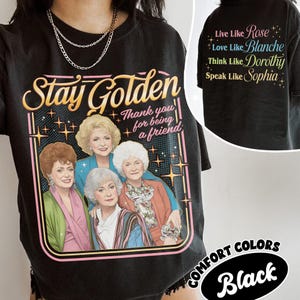 May include: Black t-shirt with a graphic featuring the phrase "Stay Golden" and an illustration of four women. The shirt also includes the text "Thank you for being a friend" and "Live Like Rose, Love Like Blanche, Think Like Dorothy, Speak Like Sophia."