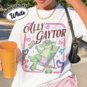 Puede incluir: Una camiseta blanca con un gráfico de un caimán verde con gafas de corazón, sentado en una silla de playa, sosteniendo una bandera arcoíris y vistiendo pantalones cortos rosas. El texto dice Ally Gaytor, rodeado de corazones y símbolos.